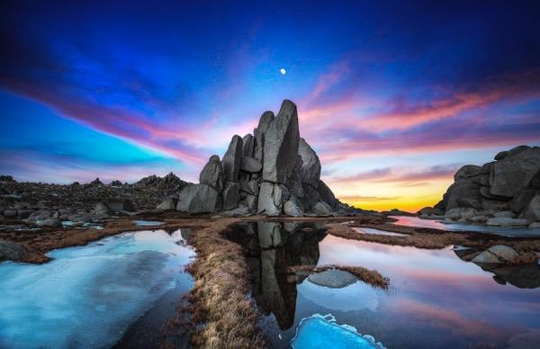 landscape with frozen lakes, browned grass and a rock mount. behind it, a blue sky with a tiny moon